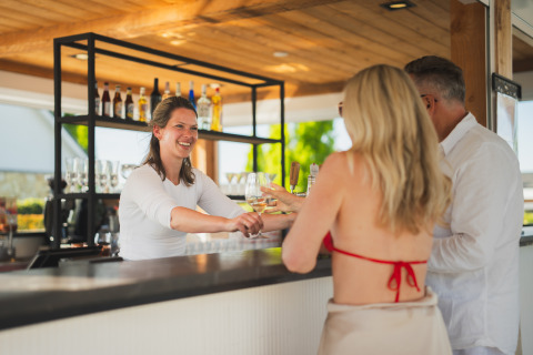 Bartender smiler og serverer drikkevarer til et par gæster ved en udendørs bar på en glampingplads.