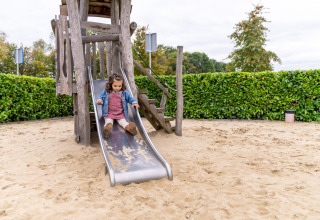 Et barn leger på en rutsjebane lavet af træ ved en glampingplads med sand og grøn hæk i baggrunden.