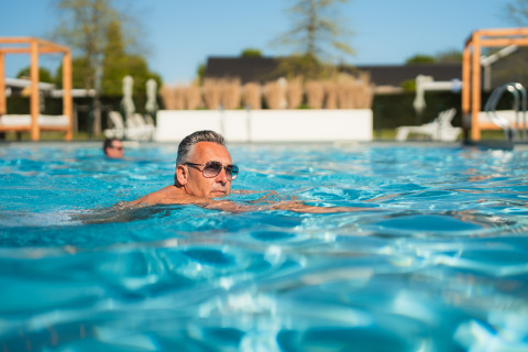 Man wearing sunglasses swimming in an outdoor glamping pool, enjoying a relaxing and sunny vacation.