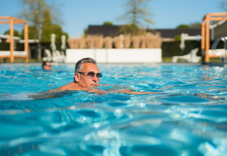 Homme avec des lunettes de soleil nage dans la piscine d'un hébergement glamping en journée ensoleillée.