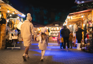 Madre e figlia camminano mano nella mano tra le bancarelle illuminate di un mercato in una struttura glamping.