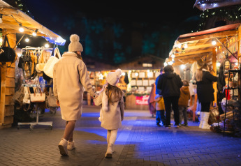 Madre e hija caminan tomadas de la mano por un mercado iluminado de noche en un alojamiento glamping.