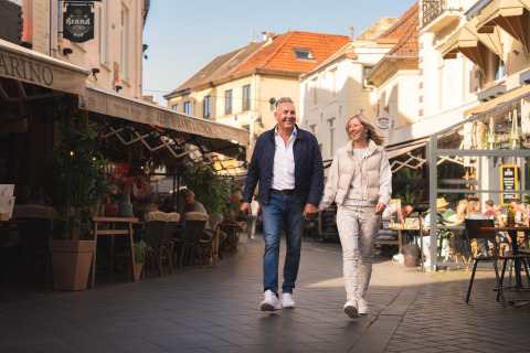 Et smilende par går hånd i hånd gennem en livlig gade med caféer og restauranter på et glampingsted.
