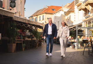 Et smilende par går hånd i hånd gennem en livlig gade med caféer og restauranter på et glampingsted.