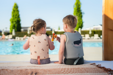 Twee kinderen met zwemvesten zitten bij een glamping-zwembad en genieten samen van een ijsje.