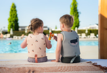 Twee kinderen met zwemvesten zitten bij een glamping-zwembad en genieten samen van een ijsje.