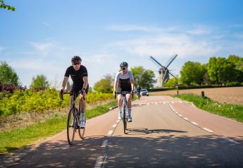 Dos ciclistas circulan por una carretera rural cerca de un molino de viento en un día soleado y claro.