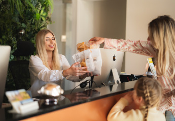 Receptionist udleverer madposer til gæst og barn ved skranken på et glampingsted, med grøn væg bagved.
