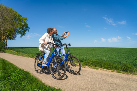 Due persone in bicicletta blu si scattano un selfie su una strada rurale accanto a campi verdi e cielo sereno.