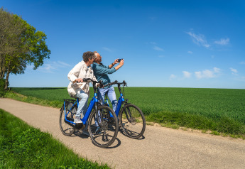 Two people on blue bikes take a selfie together on a rural road next to green fields under clear sky.
