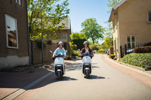 Twee mensen rijden op elektrische scooters door een zonnige woonstraat, omringd door huizen en groen.