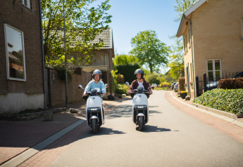 Twee mensen rijden op elektrische scooters door een zonnige woonstraat, omringd door huizen en groen.