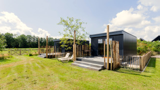 Moderne tiny house met terras, ligstoelen en houten hek in een rustige, groene landelijke omgeving.