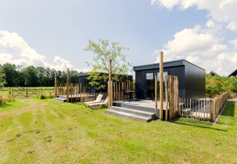Modernes Tiny House mit Terrasse, Liegestuhl und Holzzaun in einer grünen, ländlichen Umgebung.