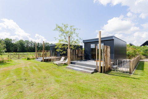 Modern tiny house with deck, lawn chairs, and wooden fence in a peaceful, green countryside setting.