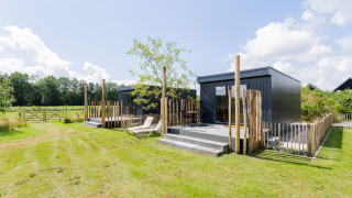 Modernes Tiny House mit Terrasse, Liegestuhl und Holzzaun in einer grünen, ländlichen Umgebung.