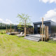 Modern tiny house with deck, lawn chairs, and wooden fence in a peaceful, green countryside setting.