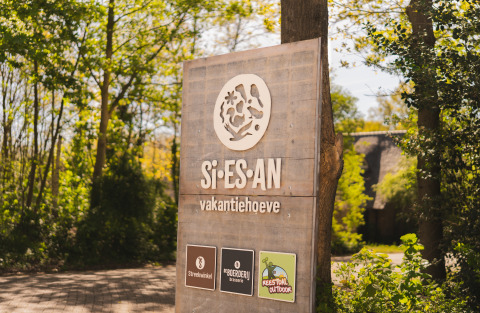 Holzschild am Eingang der Si-Es-An Glamping Unterkunft und Ferienhof, umgeben von Bäumen und Natur.