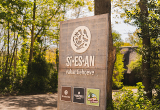 Houten bord bij de ingang van glamping en vakantiehoeve Si-Es-An, omgeven door groene natuur en bomen.