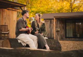 Paar sitzt auf Baumstamm am Nature house „Barn“ auf dem Campingplatz Si-Es-An, Niederlande, mit Gitarre und Wein.