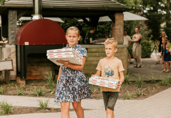 To børn bærer pizzaer ved et udendørs køkken på et glampingsted, omgivet af gæster og natur.