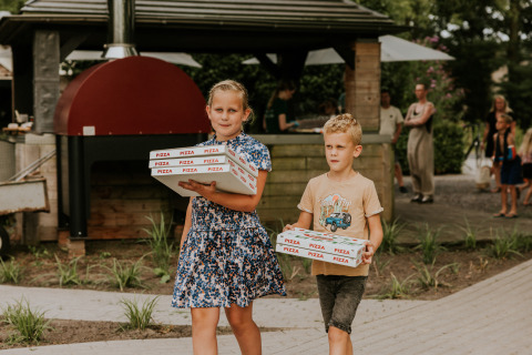 Zwei Kinder tragen Pizzakartons vor einer Outdoor-Küche an einer Glamping-Unterkunft, Gäste im Hintergrund.