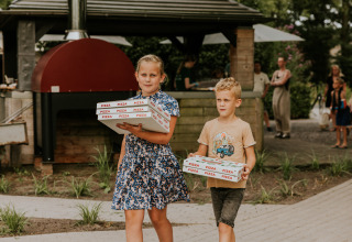 Two children carrying pizza boxes in front of an outdoor kitchen at a glamping site, with guests nearby.