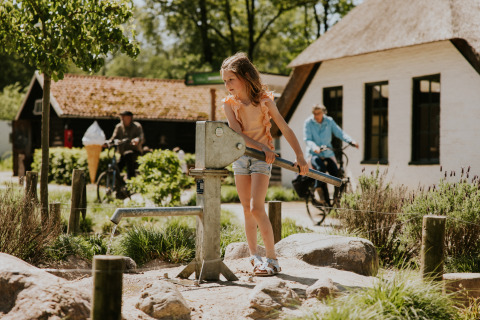Mädchen bedient eine Wasserpumpe bei einer Glamping-Unterkunft, Radfahrer und Häuser im Hintergrund.