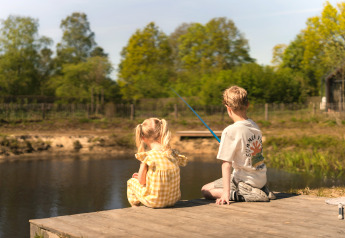 To børn sidder på en træbro og fisker ved en sø omgivet af natur ved et glamping-ophold.