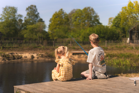 Two children sit on a wooden dock fishing by a pond, surrounded by nature at a glamping accommodation.