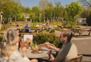 Espace de glamping extérieur avec des adultes à table et des enfants jouant en arrière-plan.