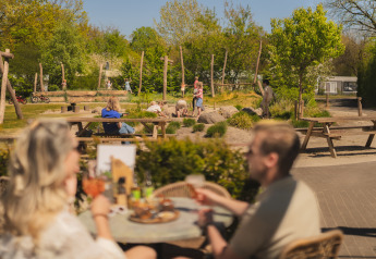 Area glamping all'aperto con adulti a tavola e bambini che giocano sullo sfondo nel parco giochi.