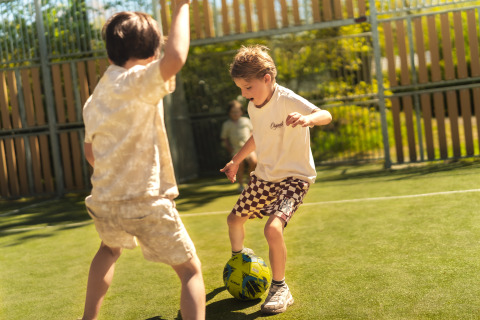 Dos niños juegan al fútbol en un alojamiento glamping, rodeados de valla y árboles en un día soleado.