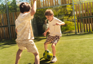 Dos niños juegan al fútbol en un alojamiento glamping, rodeados de valla y árboles en un día soleado.