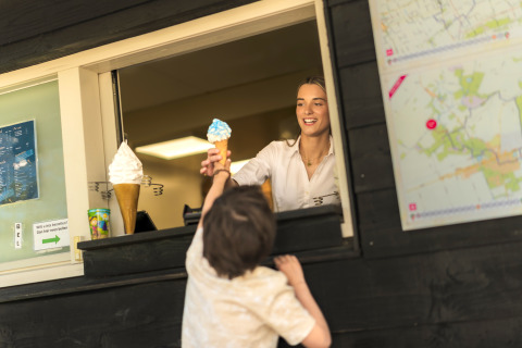 Et barn får en isvaffel fra en glad medarbejder ved isvinduet på en glampingplads, sommerstemning.