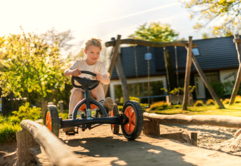 Dreng kører på pedalbil på legeplads i solskin ved glampingophold, sommerhus synligt bagved.