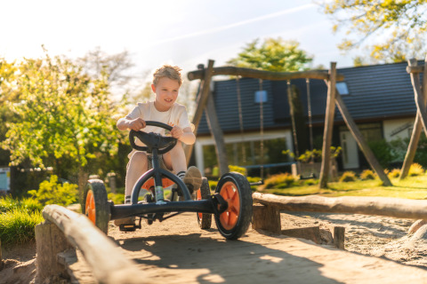 Jongen rijdt op een trapkar op de speeltuin van een glampinglocatie, met zon en huisje op de achtergrond.