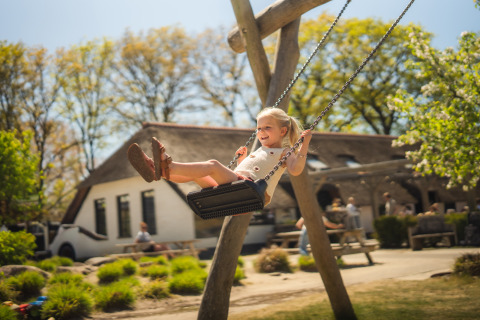 Blij meisje schommelt bij een glampingverblijf, met bomen en mensen op de achtergrond in de zon.