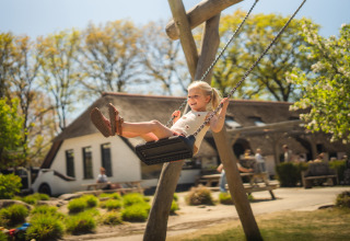 Glad pige på gynge i solskin foran et glampingopholdssted med træer og folk i baggrunden.