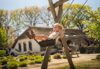 Bambina felice che si dondola all'aperto presso un glamping, con alberi e persone sullo sfondo.
