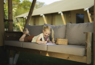 Ein kleines Mädchen malt und lächelt auf der Veranda eines Safarizeltes auf Campingplatz Si-Es-An.