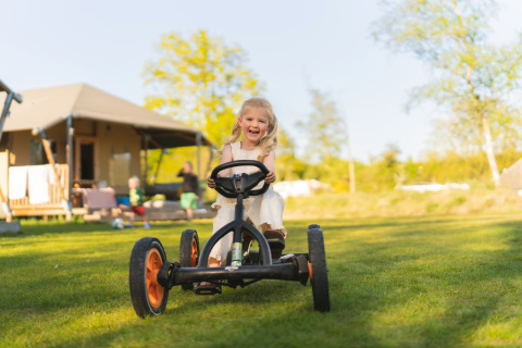 Fille heureuse sur un kart devant une tente safari au camping Si-Es-An, Pays-Bas, journée ensoleillée.