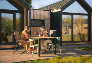 Two people share a meal outdoors at a table in front of a modern Cosy Lodge at Campsite Si-Es-An, Netherlands.