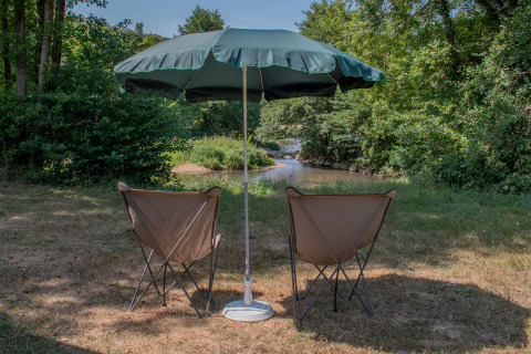 Two camping chairs under a large umbrella facing a peaceful river at a holiday park offering glamping.