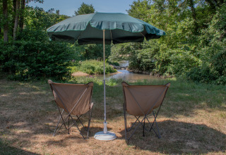 Zwei Campingstühle und ein großer Sonnenschirm stehen an einem ruhigen Fluss in einem Glamping-Park.