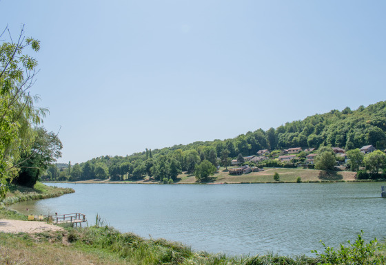 Blick auf einen See mit Wald und Ferienpark mit Glamping-Unterkünften im Hintergrund bei Sonnenschein.