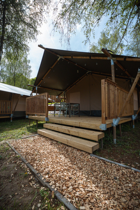 Tente safari avec terrasse en bois et chaises, située à Hameau de l'Ourthe en Belgique, entourée d'arbres.