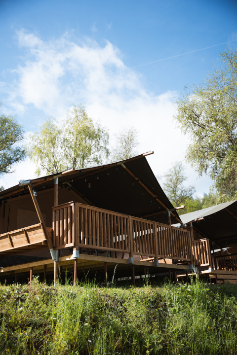 Tente safari au Hameau de l'Ourthe en Belgique avec terrasse en bois, ciel bleu et arbres en arrière-plan.