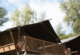 Tienda safari en Hameau de l'Ourthe en Bélgica con terraza de madera, cielo azul y árboles alrededor.