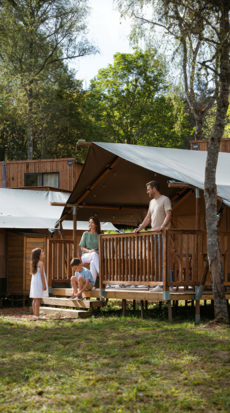 Familie nyder en solrig dag udenfor en safaritelte på Hameau de l'Ourthe i Belgien med træer omkring.